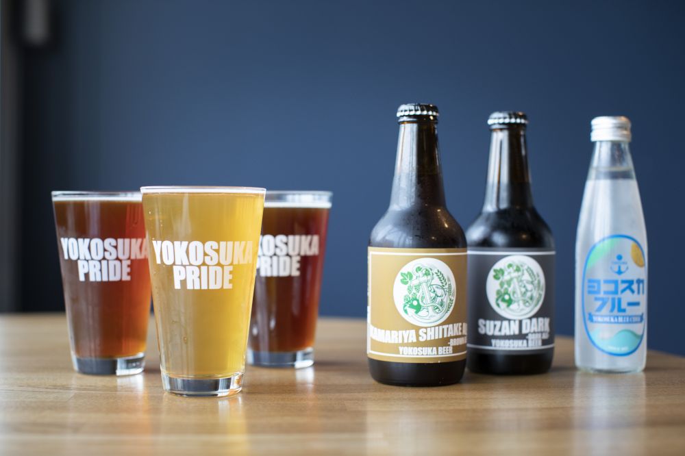 Three glasses of different craft beers on the left, and bottles of ‘Kamariya Shiitake Ale’, ‘Susan Dark’, and ‘Yokosuka Blue’ cider made with local spring water on the right at Yokosuka Beer Taproom